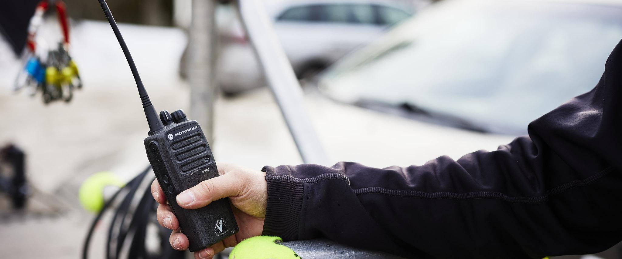 A person's hand holding a Motorla walkie talkie two-way radio on a film set with cars in the background.